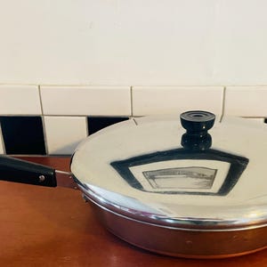 Puede incluir: Una sartén de acero inoxidable con tapa. La sartén tiene un mango negro y un pomo negro en la tapa. La sartén está sobre una superficie marrón.