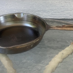 May include: A round cast iron skillet with a dark, seasoned surface, a single long handle with a hole at the end, and a small pouring spout on the rim. The metal shows signs of wear and use with a metallic sheen and dark brown tones.