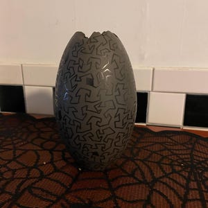 May include: A dark gray, egg-shaped decorative item with a jagged top. The surface is covered in a repeating pattern of black, abstract shapes. The item is displayed on a brown surface with a black spiderweb design.