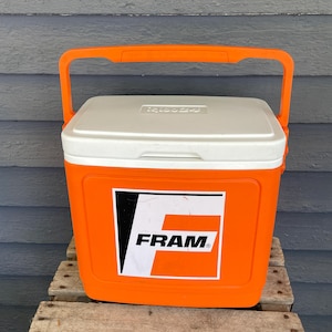 Puede incluir: Una nevera portátil Igloo naranja y blanca con un asa naranja a juego. La nevera tiene forma rectangular y presenta el logotipo de FRAM en blanco y negro en la parte delantera.