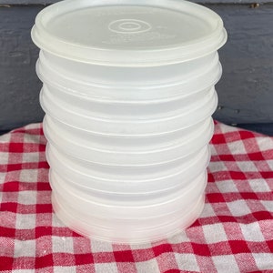Op de afbeelding: Een stapel van zeven ronde, doorschijnende, halfheldere plastic deksels voor voedselbewaarbakjes. Elk deksel heeft een cirkelvormig reliëfontwerp in het midden en het woord Tupperware is in het bovenoppervlak gegoten.