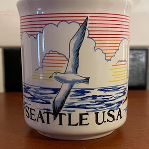 May include: White ceramic mug with a design featuring a seagull in flight over blue water. The mug has a red, orange, and yellow sky with the words "SEATTLE U.S.A." printed on the front. The mug has a white background.