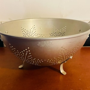 May include: A vintage aluminum colander with a wide bowl, two handles, and three curved feet. The colander has numerous small holes for draining and the brand name "MIRRO" is visible on the side. It sits on a reddish-brown surface.