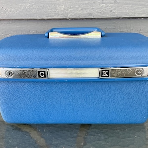 May include: A vintage blue travel case with a silver trim and handle. The case has the letters "C" and "K" on the front. The case is made of a textured material and is sitting on a glass surface.