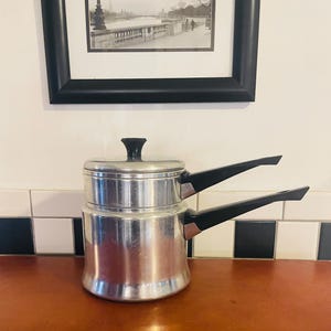 Puede incluir: Una olla doble de aluminio vintage con asas y tapa negras. La olla está sobre una superficie de madera, con un protector contra salpicaduras de azulejos en blanco y negro y una imagen enmarcada en el fondo.