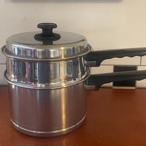 Puede incluir: Un juego de baño maría de acero inoxidable que consta de una olla inferior y un inserto superior, ambos con mangos de plástico negro con un diseño de agarre ondulado. El juego incluye una tapa de acero inoxidable con un pomo negro.