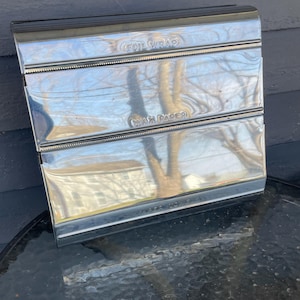 Op de afbeelding: Een vintage chromen folie- en waspapierspender. De dispenser heeft de woorden "FOIL WRAP" en "WAX PAPER" in reliëf op de voorkant. De dispenser is rechthoekig met een glanzend, reflecterend oppervlak.