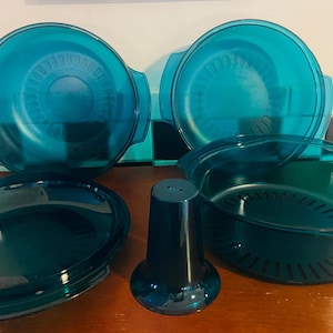 May include: A set of teal-colored kitchenware, including two round dishes with handles, a shallow oval dish, a colander-style bowl, and a tall, conical container. The items are made of a translucent material and sit on a wooden surface.