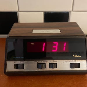 May include: A vintage digital alarm clock with a wood-grain top and black frame. The display shows "1:31 PM" in bright pink digits. It has control knobs for dimming, brightness, time set, alarm set, and alarm volume. The brand name "+Tradition" is visible.