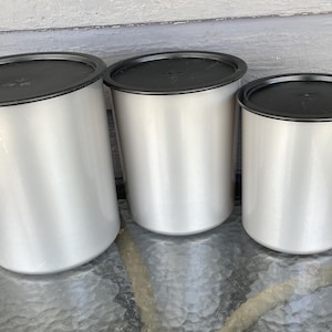 Op de afbeelding: Drie zilverkleurige canisters met zwarte deksels staan op een glazen tafel. De canisters zijn cilindrisch, met de grootste links en de kleinste rechts. De deksels lijken van matzwart materiaal te zijn.