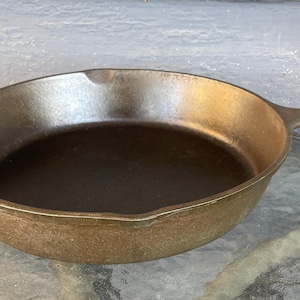 May include: A well-seasoned cast iron skillet with a long handle. The pan has a dark interior and a golden-brown exterior. The skillet is sitting on a glass table.