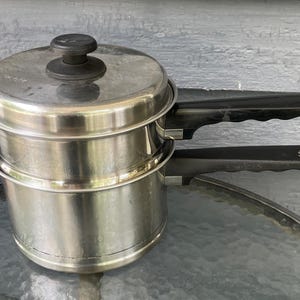 May include: A stainless steel double boiler set consisting of two stacked pots and a matching lid. Each pot features a long, black heat-resistant handle with a small white logo printed on the top. The lid has a central black knob handle.