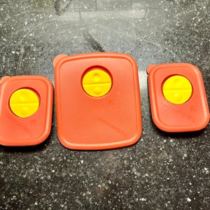 May include: Three red plastic food storage container lids with yellow buttons. The lids are labeled "Tupperware".