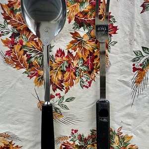 May include: A silver serving spoon and a silver fork with black plastic handles. The utensils are on a white tablecloth with a floral pattern.