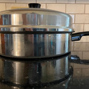 Op de afbeelding: Een gepolijste roestvrijstalen pan met bijpassend deksel en een zwarte handgreep. De pan heeft horizontale gegroefde lijnen aan de buitenzijde. Het deksel heeft een zwarte knop aan de bovenkant. Het metalen oppervlak is glad en reflecterend.
