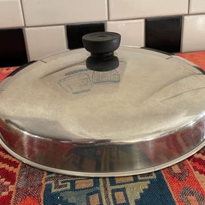 May include: A stainless steel pot lid with a black handle. The lid is round and reflective, with a slightly domed shape. The background includes a colorful patterned fabric and a black and white tiled surface.