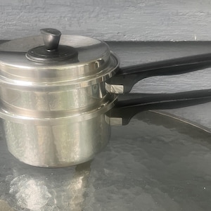 May include: A stainless steel double boiler set consisting of two stacked pots with black plastic handles and a matching lid. The top pot features a smaller diameter rim that fits securely into the base pot, both finished in a polished silver tone.