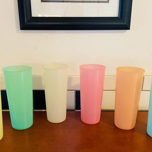 May include: Six tall, pastel-colored plastic tumblers in yellow, green, cream, pink, peach, and blue are arranged on a brown surface. The glasses are cylindrical and evenly spaced, suggesting a set for beverages.