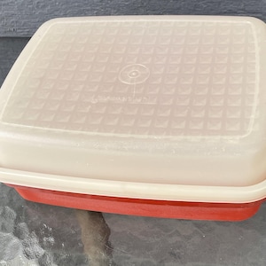 Puede incluir: Contenedor rectangular de dos tonos con tapa texturizada blanquecina y base roja. La tapa tiene un patrón de rejilla y un logotipo circular en el centro. El contenedor es probablemente para el almacenamiento de alimentos.