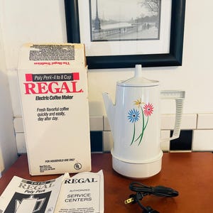 Puede incluir: Una cafetera eléctrica Regal blanca con un diseño floral, una caja a juego y un manual de usuario. La cafetera tiene una capacidad de 0,95 a 1,9 litros. La caja dice "Poly Perk".