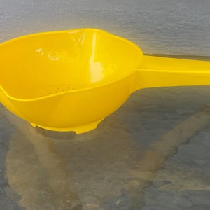 Puede incluir: Un colador de plástico amarillo brillante que presenta un cuenco ancho y redondeado con una base perforada para el drenaje y un mango largo y plano que se extiende desde un lado. El diseño incluye un pequeño orificio circular al final del mango.