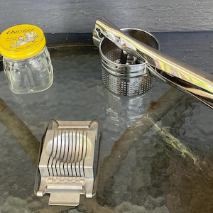 Puede incluir: Una colección de utensilios de cocina vintage sobre una mesa de cristal. Incluye un tarro de cristal con tapa amarilla, un prensador de patatas cromado y un cortador de huevos. La tapa del tarro tiene la palabra "Osterizer".