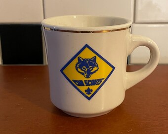 Puede incluir: Taza de cerámica color crema con borde dorado y asa. La taza presenta un logotipo azul y amarillo de los Cub Scouts con la cabeza de un lobo y las palabras "Cub Scouts". Una flor de lis en la parte inferior.