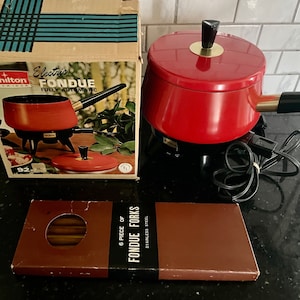 Könnte beinhalten: Ein vintage roter elektrischer Fonduetopf mit schwarzem Sockel und goldenem Knopf auf dem Deckel. Der Topf steht auf einer schwarzen Arbeitsplatte und ist eingesteckt. Zum Topf gehört eine Schachtel mit sechs Fonduegabeln.