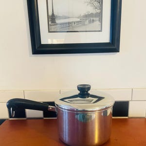 May include: A stainless steel saucepan with a black handle and lid. The pot sits on a brown surface, with a black and white tiled backsplash. A framed black and white print hangs above the pot.