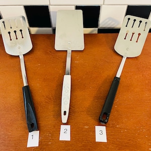 May include: Three stainless steel spatulas with varying handle designs. Two have black handles, and one has a white handle with a floral pattern. The spatula heads have different shapes, including slotted and solid designs. Kitchen utensils.