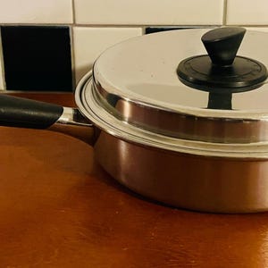 Puede incluir: Una cacerola de acero inoxidable con tapa y un mango largo negro. La tapa tiene un pomo negro. La cacerola está sobre una superficie marrón con un fondo de azulejos blancos y negros.