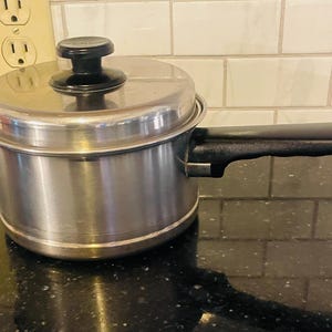 Puede incluir: Un cazo de acero inoxidable con tapa a juego y un mango largo de color negro. La olla presenta un cuerpo cilíndrico con acabado metálico pulido, una tapa plana con un pomo central negro y un mango negro texturizado que sale del lateral.