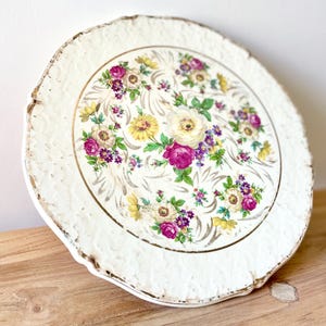 Può includere: Un piatto rotondo in ceramica con un bordo smerlato e testurizzato color crema con accenti dorati. Il centro presenta un motivo floreale colorato con fiori rosa, gialli e viola, foglie verdi e volute dorate su uno sfondo bianco.