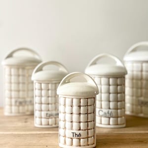 Puede incluir: Un conjunto de botes de cerámica blanca con un patrón de cuadrícula y asas arqueadas. Los botes están etiquetados con las palabras "Thé" y "Café". Los botes están sobre una superficie de madera.