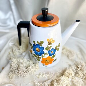 Könnte beinhalten: Weiße Emaille-Teekanne mit schwarzem Griff und Deckel sowie einem orangefarbenen Rand. Die Teekanne ist mit blauen und orangenen Blumenmustern verziert. Der Ausguss ist weiß. Die Teekanne steht auf einer weißen Oberfläche.