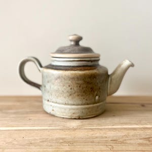 Peut inclure: Une théière en céramique avec un couvercle, une anse et un bec verseur. La théière est principalement beige avec des accents et des mouchetures marron. Elle repose sur une surface en bois. La théière a un design classique, pour servir le thé.