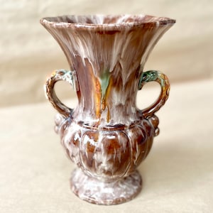 Peut inclure: Un vase en céramique avec une finition émaillée coulante brune et blanche. Le récipient présente un bord large et évasé, un corps bulbeux avec des cannelures verticales et deux anses incurvées ornées de chaque côté du col.