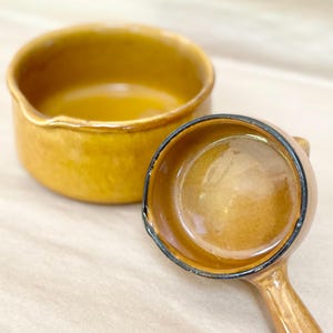 Peut inclure: Une paire de récipients de cuisine en céramique émaillée d'une teinte jaune moutarde chaude. L'un est un bol rond avec un petit bec verseur, tandis que l'autre est une petite poêle circulaire à manche avec un bord sombre et contrasté.