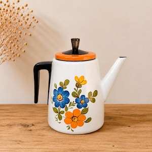 Peut inclure: Théière en émail blanc avec une poignée et un bec noirs, et un couvercle orange. La théière est décorée de motifs floraux bleus et oranges. La théière est posée sur une surface en bois.
