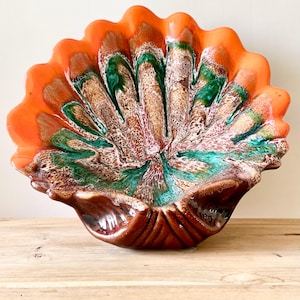 Peut inclure: Un plat de service décoratif en céramique avec un bord festonné. Le plat présente une palette de couleurs vibrantes d'orange, de vert, de marron et de blanc. Le plat est posé sur une surface en bois.
