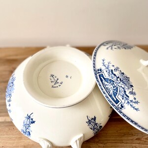 Peut inclure: Un plat de service en céramique blanche avec un couvercle, orné de motifs floraux bleus. Le plat a des poignées et une base surélevée. Le couvercle est partiellement ouvert, révélant l'intérieur.