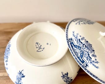 Op de afbeelding: Een witte keramische serveerschaal met deksel, versierd met blauwe bloempatronen. De schaal heeft handvatten en een verhoogde voet. Het deksel is gedeeltelijk open en onthult de binnenkant.