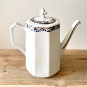 Peut inclure: Théière en céramique blanche de forme géométrique. Le couvercle et le bord présentent une bordure décorative avec des accents bleus, jaunes et dorés. La théière a une anse et un bec avec une garniture dorée. Elle repose sur une surface en bois.