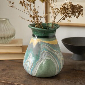 Peut inclure: Un vase décoratif en céramique avec un bord vert et un motif marbré dans les tons de vert, gris, jaune et bleu. Le vase contient des fleurs séchées. Le vase mesure environ 20 cm de haut.
