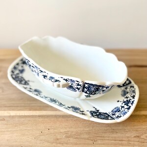 Peut inclure: Un saucier et sa soucoupe en céramique blanche avec un motif floral et des volutes bleues. Le saucier a un bord festonné. La soucoupe est rectangulaire avec un bord incurvé. L'ensemble est sur une surface en bois.