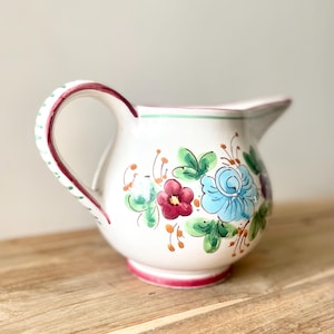 Peut inclure: Pichet en céramique blanche avec un corps arrondi et une anse. Le pichet est décoré de fleurs peintes à la main dans des tons de bleu, de rouge et de violet, avec des feuilles vertes. La anse et la base sont bordées de rouge, et le bord a une bande verte.
