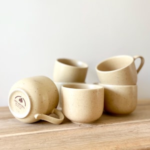 Peut inclure: Un ensemble de cinq tasses en céramique beige clair avec une finition mouchetée. Une tasse est posée sur le côté, révélant un tampon circulaire sur la base avec l'inscription GRES NATUREL et une icône d'arbre. Les tasses ont des anses.