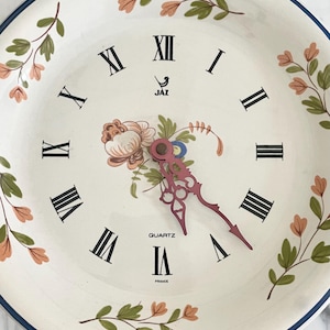 Peut inclure: Une horloge à quartz blanche avec des chiffres romains noirs et un motif floral. Le cadran de l'horloge présente un arrangement floral rose et vert au centre, avec des aiguilles roses. L'horloge est marquée "JAZ" et "France".