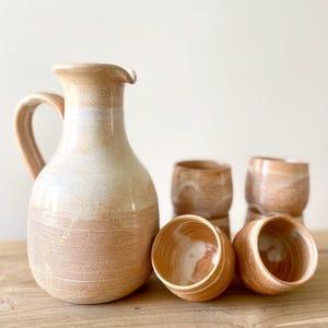 May include: A ceramic pitcher with a handle and four matching cups, all featuring a light beige and tan glaze. The pitcher has a rounded body with fine horizontal ridges on the lower half, while the cups have a smooth interior and textured exterior.