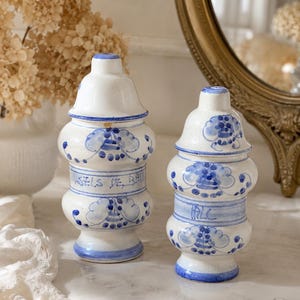Peut inclure: Deux pots en céramique blancs avec des motifs floraux bleus et des couvercles. Les pots ont une forme arrondie avec une bande bleue et le texte "SELS DE BAIN" sur l'un. Les couvercles sont blancs avec des détails bleus.